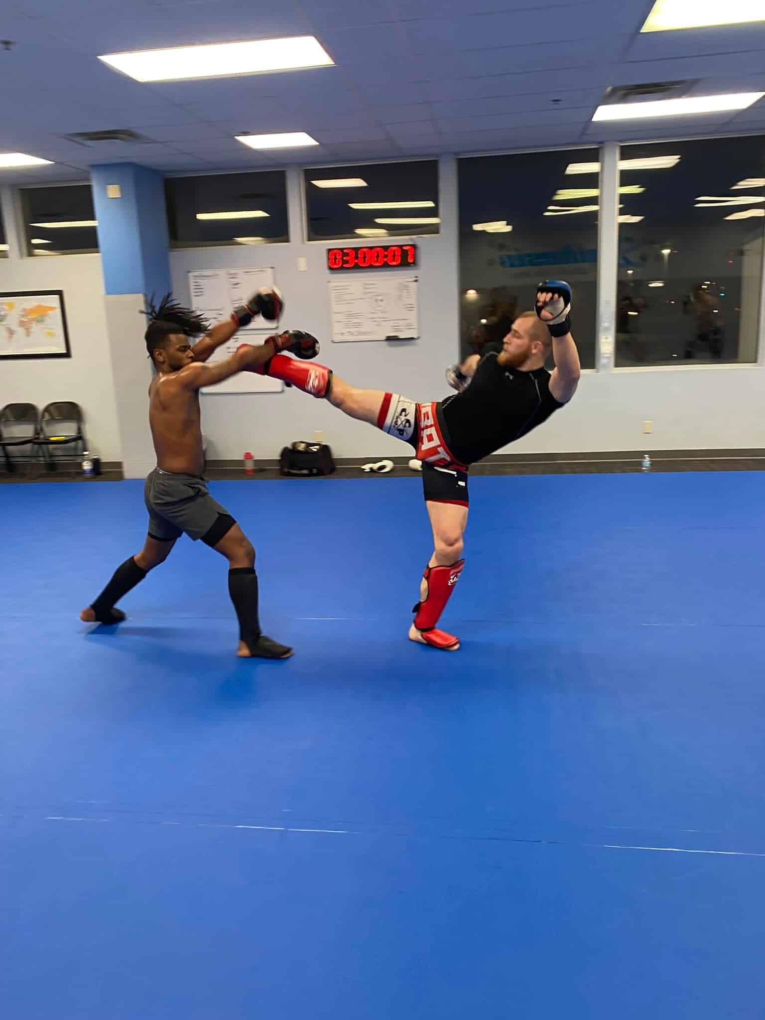 two members in sparring equipment spar on the mat, one kicks high and one blocks